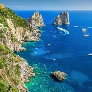 Una costa rocciosa e verdeggiante con edifici si affaccia su un mare blu profondo punteggiato di barche vicino a due grandi faraglioni.