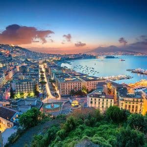 Veduta aerea di una città costiera illuminata al crepuscolo, con un porto pieno di barche e una grande montagna sullo sfondo.