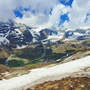 Una vista panoramica di una catena montuosa innevata su una valle verde con un piccolo villaggio, vista da un pendio innevato sotto un cielo nuvoloso.