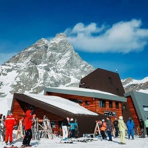 Eine WeRoad-Gruppenreise mit Skiern und Snowboards versammelt sich vor einer Berghütte, im Hintergrund ein großer, schneebedeckter Gipfel unter blauem Himmel.