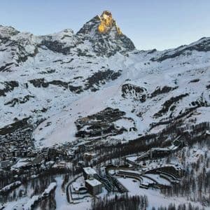Una vista aerea di un villaggio sciistico incastonato in una valle innevata ai piedi di una grande vetta montuosa illuminata dal sole.