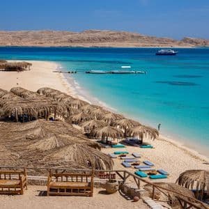 Vue plongeante sur une plage tropicale, avec des parasols en paille le long du rivage aux eaux turquoise cristallines et des collines désertiques en arrière-plan.
