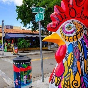 Una grande statua di gallo, dipinta a colori vivaci, si trova su un marciapiede cittadino accanto a un segnale stradale sotto un cielo sereno.