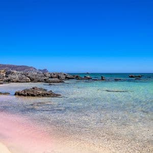 Eine Küste mit auffällig rosafarbenem Sand trifft auf klares, flaches türkisfarbenes Wasser, dazu dunkle Felsen und ferne Hügel unter einem strahlend blauen Himmel.