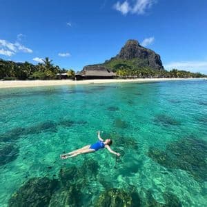 Una mujer en traje de baño azul flota de espaldas en agua clara turquesa cerca de una playa tropical con una montaña al fondo.