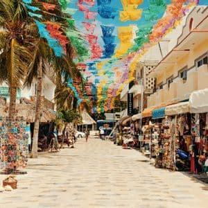 Una strada lastricata fiancheggiata da negozi e palme è decorata con bandiere di carta colorate appese in alto.