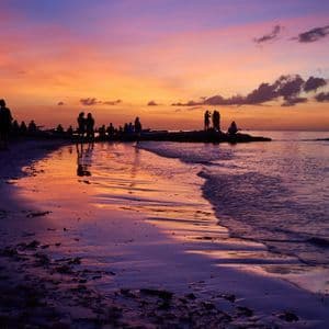 Sagome di un viaggio di gruppo WeRoad su una spiaggia durante un tramonto vibrante, con cieli viola e arancioni che si riflettono sulla sabbia bagnata.