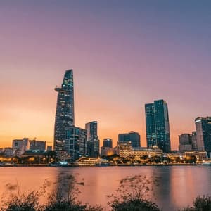 A modern city skyline with illuminated skyscrapers reflecting in a river against a colorful sunset sky.