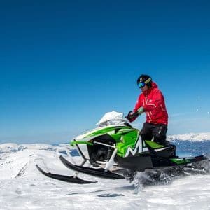Una persona con una chaqueta roja conduce una moto de nieve verde por una cordillera nevada bajo un cielo azul claro.