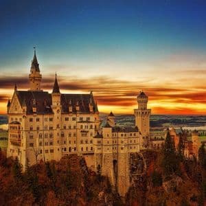 A large castle with multiple towers sits on a forested cliff, overlooking a valley with lakes under a colorful sunset sky.
