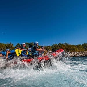 A WeRoad group trip paddles a red raft through splashing whitewater rapids under a clear blue sky.