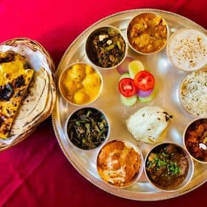 Veduta dall'alto di un thali indiano con diversi curry e riso, accompagnato da un cestino di pane piatto su un panno rosso.