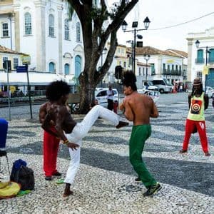 Eine Gruppe von Männern führt Capoeira, eine brasilianische Kampfkunst, auf einem Kopfsteinpflasterplatz auf, der von historischen Gebäuden umgeben ist.