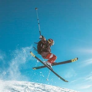 Uno sciatore a mezz'aria tiene un selfie stick mentre salta su una montagna innevata sotto un cielo azzurro e limpido.