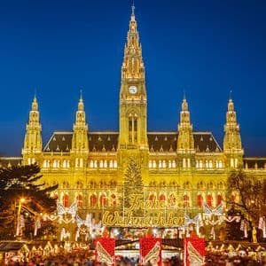 A large, illuminated building with a clock tower at night behind a bustling Christmas market with festive lights and a tree.
