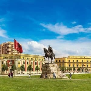 Una gran estatua ecuestre en una plaza de la ciudad con una bandera albanesa ondeando frente a edificios históricos y una mezquita en un día soleado.