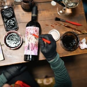 Vista dall'alto di una persona con guanti neri che dipinge l'etichetta di una bottiglia di vino su un tavolo di legno, circondata da materiali d'arte.