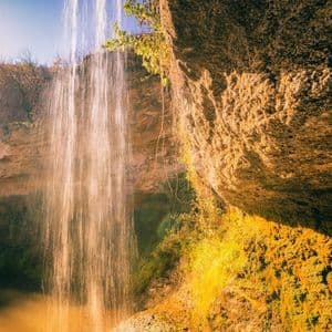 Una cortina d'acqua di una cascata è illuminata dalla luce dorata del sole mentre scende lungo una parete rocciosa muschiosa.
