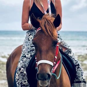 Una mujer de un viaje en grupo de WeRoad montando un caballo marrón en una playa, con el mar de fondo.