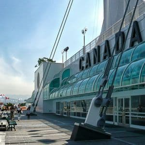 Una bandiera canadese sventola su un molo sul lungomare accanto a un edificio moderno con la scritta 'CANADA' sulla sua facciata sotto un cielo soleggiato.