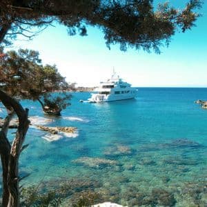 Un grande yacht bianco è ancorato in una baia calma, limpida e turchese, visto da una costa rocciosa sotto i rami degli alberi.
