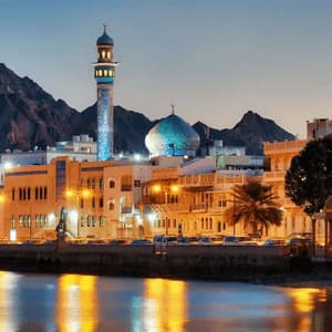 An illuminated mosque with a blue tiled dome and minaret on a city waterfront at dusk, with mountains in the background.