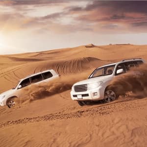 Two white SUVs kick up sand while driving down a steep dune in the desert at sunset.