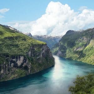 A turquoise fjord winds between steep, forest-covered mountains with snow-capped peaks under a blue sky with white clouds.