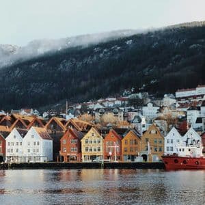 A row of colorful wooden houses lines a waterfront, with a red boat moored alongside and a forested hill rising in the background.