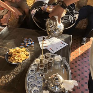 A person pours tea from an ornate silver teapot into small glasses for a WeRoad group trip, with cookies on the table.