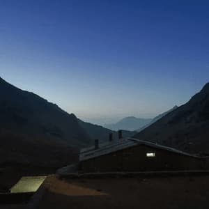 A mountain refuge with a single glowing window and a lit tent nestled in a dark valley at dusk.