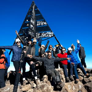 Un voyage de groupe WeRoad posant pour une photo sur un sommet de montagne rocheux, à côté d'une grande borne triangulaire.