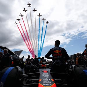 From behind a Formula 1 car, a driver and crew watch jets perform a flyover, leaving trails of red, white, and blue smoke.