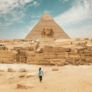 Une personne marche sur le sable vers le Grand Sphinx, avec une grande pyramide en arrière-plan sous un ciel nuageux.