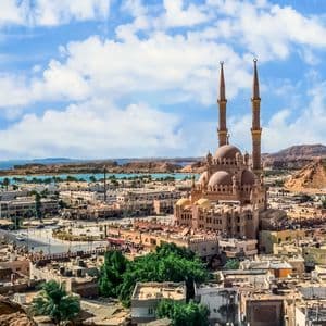 Vue aérienne d'une ville avec une grande mosquée et ses deux minarets, bordée par des eaux bleues et des collines arides sous un ciel nuageux.