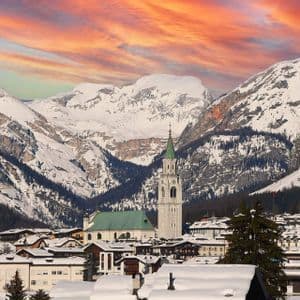 Un pueblo cubierto de nieve con un alto campanario de iglesia nestled contra montañas nevadas bajo un cielo naranja y rosado.