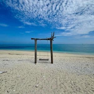 Un'altalena rustica in legno, vuota, su una spiaggia sabbiosa, di fronte all'oceano calmo e blu sotto un cielo parzialmente nuvoloso.