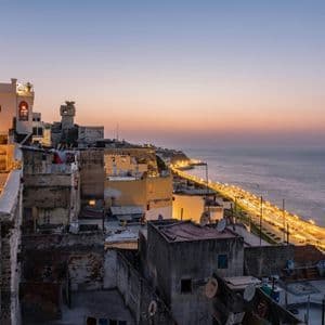 Una vista sobre los tejados de una ciudad costera histórica al atardecer, con una carretera iluminada que bordea el mar.