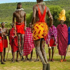 Due uomini Maasai in abiti tradizionali saltano in alto durante una danza cerimoniale, mentre altri uomini osservano su una pianura erbosa.