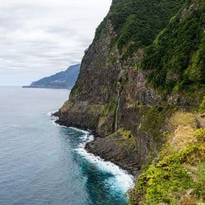 Ein Wasserfall stürzt eine steile, grün bewachsene Felswand hinunter ins Meer, wobei Wellen gegen die felsige Küstenlinie schlagen.