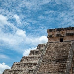 Una vista dal basso di un'antica piramide a gradoni in pietra che si eleva verso un cielo blu con nuvole bianche sparse.