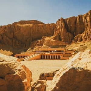Un grande tempio terrazzato costruito alla base di un'imponente scogliera rocciosa, visto da tra due grandi massi sotto un cielo limpido.