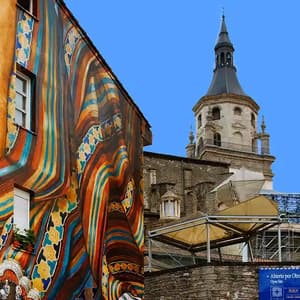 Un edificio con un grande murale colorato si erge accanto a un'antica torre campanaria in pietra sotto un cielo azzurro e limpido.