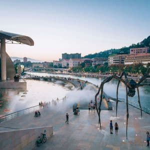Una grande scultura di ragno in metallo su una passeggiata lungofiume accanto a un edificio moderno al crepuscolo, con persone che camminano nella nebbia.