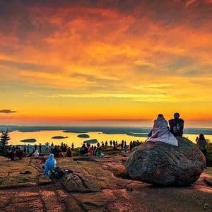 Una folla di persone su una vetta rocciosa osserva un tramonto vivace, arancione e giallo, su uno specchio d'acqua costellato di isole.