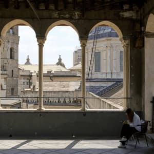 Una donna è seduta su una sedia su un balcone ombreggiato con archi in pietra, che incorniciano una vista sui tetti della città e una grande cupola.