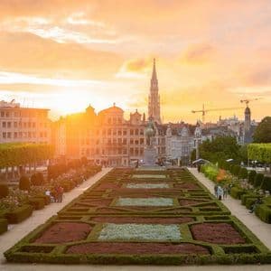 Un giardino formale con siepi geometriche e uno skyline cittadino con un campanile alto sullo sfondo durante un tramonto dorato.