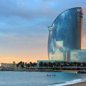 A modern, sail-shaped glass building stands on a beach next to the ocean at sunset, with palm trees lining the shore.