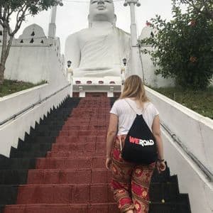 A person with a WeRoad backpack walks barefoot up a long staircase towards a large, white seated Buddha statue.