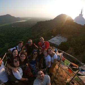 Eine WeRoad-Gruppe macht ein Selfie auf einem Berggipfel bei Sonnenaufgang, mit einem weißen Stupa und einem üppigen Tal im Hintergrund.